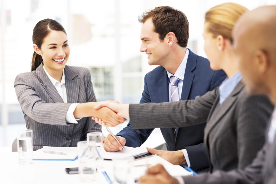 Happy young businesswomen shaking hands while sitting with multi-ethnic colleagues in meeting. Horizontal shot.
